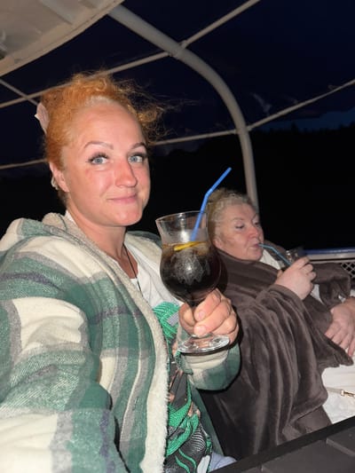 The image depicts two women on what appears to be a boat at night. The woman in the foreground, with red curly hair and blue eyes, is holding a glass with a dark beverage and a lemon slice. She is wearing a green and white plaid jacket. Behind her, another woman with blonde curly hair is partially visible, also holding a drink and wrapped in a brown blanket. They are seated under a dark blue canopy with white structural elements, suggesting an outdoor setting. The background is dark, with faint outlines of trees visible against the night sky. The location is identified as Makarovo, Russia.