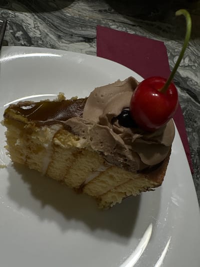 A slice of chocolate cake with whipped cream and a cherry on top sits on a white plate. The cake has layers of sponge cake and cream, with a glossy chocolate topping. A single red cherry with a green stem rests on the whipped cream. The plate is on a dark, marbled surface. A maroon napkin is partially visible to the right of the plate. The image is well-lit, highlighting the textures and colors of the dessert. The location is specified as the city of Dalgopol, Bulgaria.
