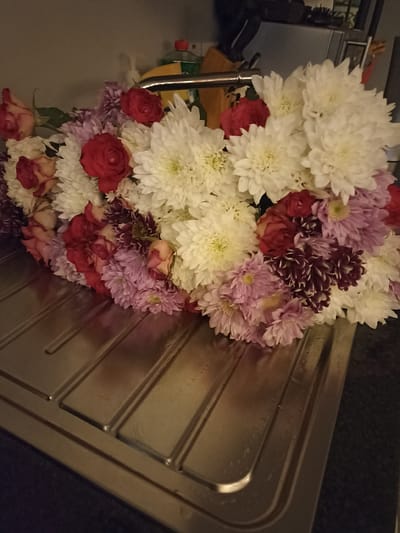A bouquet of flowers, predominantly white chrysanthemums with accents of red roses, pink and dark purple dahlias, and some lighter pink flowers, rests on a stainless steel sink. The sink has visible ridges and appears wet, with droplets of water present. In the background, a kitchen sink faucet is visible, along with a yellow bottle of cleaning product with a red cap, and a wooden cutting board. The overall setting suggests a domestic kitchen environment. The image does not contain information about people, actions, interactions, or a specific location in Annagry, Ireland.