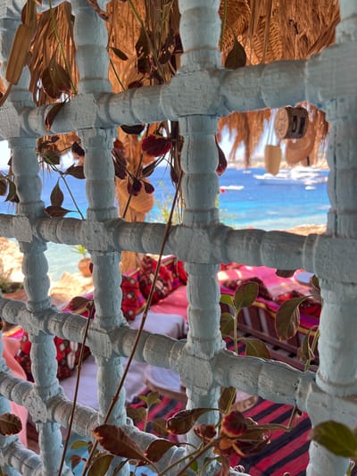 An eye-level, medium shot captures a view through a rustic, light blue, wooden lattice structure, possibly a balcony railing or screen, at an outdoor seating area in Sharm El Sheikh, Egypt. The lattice is weathered and textured, with some of the paint chipped. Several thin, dry branches with dark leaves hang in front of and through the lattice, adding a natural, somewhat overgrown feel to the scene.
Beyond the lattice, the image opens up to a vibrant seascape. The water is a brilliant turquoise and blue, stretching out to the horizon under a bright, sunny sky. Several boats, including a large white yacht, are visible on the water, indicating a popular tourist or leisure destination. In the foreground, beneath the lattice and on a lower level, there appears to be a seating area with cushions and textiles. These textiles display a mix of colors and patterns, with prominent stripes of red, black, and possibly pink, along with some richly patterned cushions that suggest a bohemian or traditional aesthetic. A thatched roof or canopy is visible above, providing shade. The overall impression is one of a relaxed, scenic spot by the sea, likely a café or restaurant, in a resort town like Sharm El Sheikh. There are no people directly visible in the frame, but the setup implies a space for human use and enjoyment.