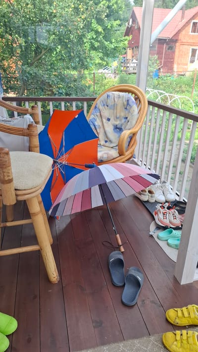 The image depicts an outdoor porch with wooden flooring. On the porch, there are two open umbrellas, one with blue and orange panels and another with multi-colored pastel stripes. Several pairs of shoes are visible: two pairs of white sneakers, one pair of beige casual shoes, a pair of pink and white sneakers, a pair of light blue clogs, and two yellow shoes. Two dark gray slip-on sandals are also present. To the left, a section of a wooden chair with a light-colored cushioned seat is visible. In the background, a rattan chair with a patterned cushion is placed on the porch. Beyond the porch railing, lush green trees and a red wooden house with a pitched roof are visible. The scene suggests a domestic setting, possibly a porch or veranda of a dacha (summer house) in a rural or suburban area, consistent with the provided location of Орбита Снт (терпигорьево), Russia.