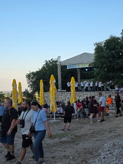 A crowded outdoor event is underway on a rocky hillside at dusk. A stage, adorned with a large tan tent and a banner reading "ФОЛКЛОРЕН ФЕСТИВАЛ" (Folklore Festival), is set up at the top of the hill, featuring a band in white uniforms performing with brass instruments. Below the stage, a crowd of people are gathered, many seated on stone benches or standing. Tall, yellow umbrellas are lined up in front of the stone wall of the stage area, some open and some closed. In the foreground, several individuals are walking along a gravel path, with more people visible in the middle ground, some seated and some standing. The sky is a clear, pale blue, suggesting the late evening. Trees and foliage frame the right side of the scene, and a distant landscape is visible in the background. The location is identified as Gara Oreshets, Bulgaria.