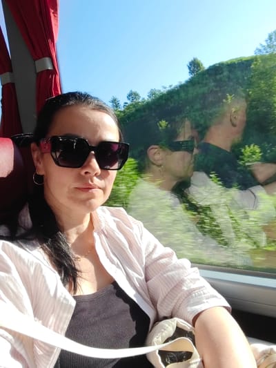 This is a candid, selfie-style photo taken from inside a bus, likely in Buknari, Georgia. The main subject is a woman with dark hair, wearing large, black, square-framed sunglasses and a light pink and white striped shirt over a dark gray ribbed tank top. She has a thin gold necklace with a small pendant. She appears to be looking slightly to her left, with a neutral expression.
The setting is a bus interior, indicated by the red seat on the left and the large window on the right. Through the window, a reflection shows another person, possibly a man, in the seat behind her, wearing a dark shirt. Outside the window, lush green trees and foliage are visible against a clear blue sky, suggesting it is a sunny day. The trees are dense and appear to be on a hillside. The motion blur visible in the reflection and the scenery outside suggests the bus is in motion.
The time of day appears to be daytime, given the bright sunlight and blue sky. There is no visible text in the image. The overall mood is relaxed and casual, possibly during a scenic journey.