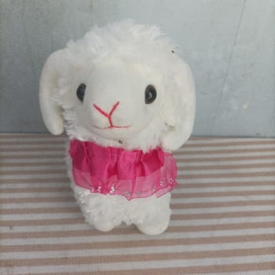 A white plush toy, resembling a lamb or sheep, stands centrally on a striped surface. The toy features black eyes and a red stitched nose/mouth forming a 'Y' shape. It wears a fuchsia pink ruffled collar or skirt-like accessory, embellished with silver glitter, around its upper body. Several small, dark insects are present on the plush toy, with one visible on the top of its head and another on its lower face. The toy rests on a surface with alternating light brown and off-white horizontal stripes. The background consists of a plain, light grey wall. The scene is located in Pasir Muncang, Indonesia.