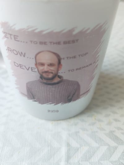 A close-up, high-angle shot shows a white ceramic mug with a printed design. The design features a man's portrait and text. The man, with a shaved head and a beard, is wearing a gray, ribbed turtleneck sweater. The text around the man's portrait reads, "ETE... TO BE THE BEST," "ROW... TH THE TOP," and "DEVE... TO REMAIN A". Below the portrait, the year "2020" is printed in dark gray. The mug is placed on a white tablecloth with a geometric pattern. The image does not provide any information or visual cues about the city of Oława, Poland.