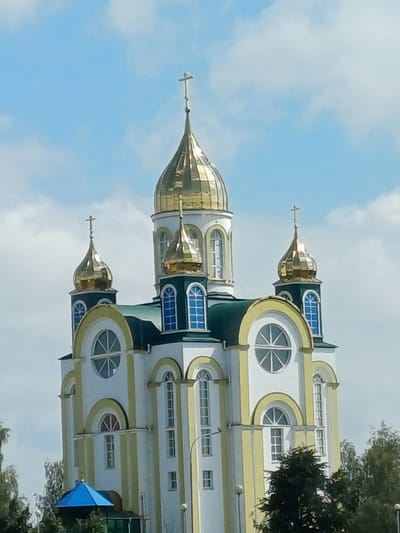 The image shows the Ascension Cathedral in Kobryn, Belarus, on a sunny day with a partly cloudy sky. The cathedral is a prominent building with multiple golden domes, white walls, and yellow decorative trim. It features arched windows with blue glass and decorative circular windows. The building is surrounded by trees and greenery.