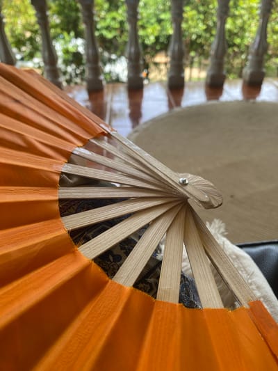 A close-up shot depicts an open hand fan with orange fabric and light-colored wooden slats. The central pivot point of the fan is visible, held together by a small metal fastener. The fan partially obscures a background with blurred elements including what appears to be foliage seen through blinds. The location is Baan Nai Suan, Thailand.