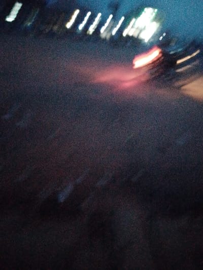 The image appears to be a nighttime scene in Urualla, Nigeria, possibly taken with motion blur. The prominent features are blurred streaks of light, likely from streetlights and vehicle headlights/taillights. The road surface is dark and indistinct. Due to the blur, identifying specific people, subjects, or objects is difficult. Actions and interactions are undiscernible. The environment seems to be urban, suggested by the presence of street lighting.