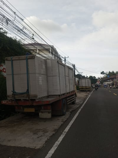 Two trucks laden with stacks of gray blocks, secured by straps, are parked beside a road. The foreground truck is mostly visible, while the second one is partially obscured. The road has a white line marking its edge. Overhead power lines crisscross the sky. A building and trees are also visible in the background, with light traffic on the road. The sky is overcast.