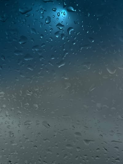 The image depicts rain droplets on a glass surface. The left side of the image is dominated by a blue hue, with more prominent and well-defined droplets. The right side transitions to a gray tone with a high density of droplets. There is no indication of the image location being in Arandjelovac Municipality, Serbia, and there are no people, objects, actions, or interactions evident in the frame.