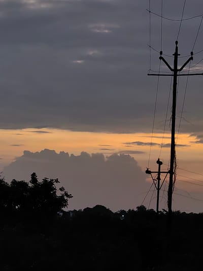 A silhouette of a power line pole with wires is positioned on the right side of the frame, against a twilight sky. The sky is a gradient of dark gray at the top, transitioning to a bright orange and yellow band in the middle, with darker clouds below it. The bottom of the image is a dark silhouette of trees and vegetation, with a faint indication of distant lights and structures. The scene appears to be an outdoor, rural or semi-rural environment in Dattapur, India, during dusk or dawn. There are no people visible in the image.
