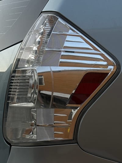 The image shows a close-up of a car's taillight, specifically the red portion and the clear plastic housing. The taillight is mounted on the body of a silver car. The housing has various textured and ribbed sections, designed to refract and direct light. The red brake light area is visible within the clear housing. Reflections from the surrounding environment are prominent on the smooth, clear plastic. These reflections show a building with brown walls and windows, suggesting an urban or residential setting. The lighting indicates it is daytime, likely with bright sunlight, given the strong reflections. There are no people visible, and the focus is solely on the automotive component and its reflections. The location is specified as Mastaga, Azerbaijan, but there are no specific visual cues in the image to confirm this without the provided context. The overall impression is of a clean, well-maintained vehicle component.