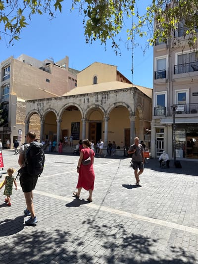 A sunny daytime scene unfolds in a public square paved with light gray stone tiles, likely in Heraklion, Greece. A large building with arched walkways and a pale yellow facade dominates the background, suggesting a historical or religious structure. Adjacent to it is a modern, multi-story building with balconies and windows.

In the foreground, several individuals are navigating the square. A man with a large black backpack is walking away from the viewer, accompanied by a young child in a floral dress. To their right, a woman in a vibrant red dress walks past. Further to the right, a man in a grey t-shirt and shorts walks towards the viewer, carrying a shopping bag. Near the building on the right, an elderly person dressed in white is seated on a stool, appearing to be a street performer.

The square is shaded in parts by the foliage of trees overhead, casting dappled patterns on the ground. Signs are visible on some of the storefronts, including one for "MARKASI OPTIKA KAO", indicating shops are present. The overall atmosphere suggests a lively urban environment during daylight hours.