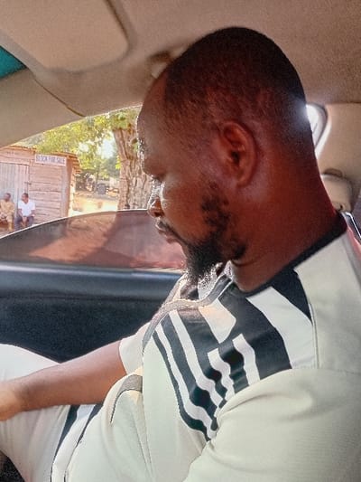 A man with dark skin and a short beard is looking down. He's wearing a white shirt with black and white stripes on the upper portion. The image seems to be taken from inside a vehicle, as parts of the interior are visible. Outside the window, a wooden structure with "BLOCK FOR SALE" written on it is seen, along with a few people gathered nearby. The lighting suggests it could be daytime.