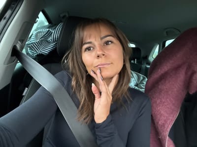 An indoor photograph captures the interior of a vehicle, in a location within Portes-lès-Valence, France. A female subject occupies the front passenger seat, secured by a dark gray seatbelt. She has brown hair with bangs and is wearing a dark, long-sleeved top. Her right hand, adorned with dark fingernail polish, rests on her chin, and her gaze is directed towards the vehicle's front-left side (viewer's right). An "AIRBAG" label is visible on the pillar next to her. In the rear, a second individual is partially visible behind the primary subject, wearing a light gray hooded garment. To the far right of the frame, a third individual is partially obscured, identifiable by a maroon-colored, textured jacket. A striped garment is present on a rear seat headrest. The exterior environment, seen through the vehicle's windows, appears overcast and indistinct.