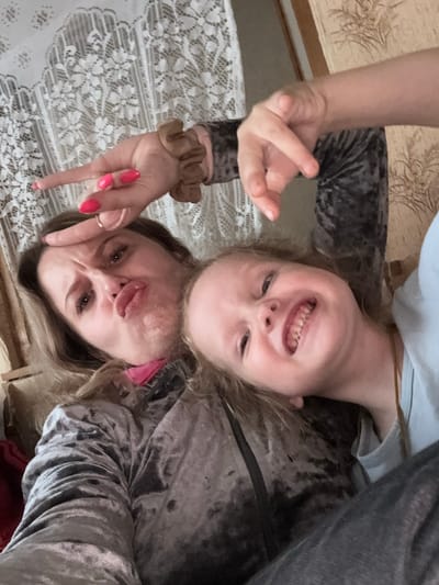 In an indoor setting in Severnyy, Russia, a woman and a young child are captured in a close-up selfie. The woman, with blonde hair and vibrant pink fingernails, is making a duck face and has her index and middle fingers raised in a peace sign, her ring finger adorned with a simple band. She wears a dark, plush velvet top with a leopard-like pattern. To her right, the child, with blonde hair and a playful expression, bares their teeth in a wide smile. The child is dressed in a light blue t-shirt. The background reveals a lace curtain to the left and a textured fabric, possibly upholstery, to the right. The overall scene conveys a moment of shared joy and lightheartedness.