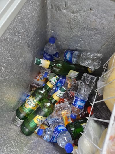 The image displays the interior of a chest freezer or large cooler, characterized by its textured, metallic-gray walls. The space is filled with a variety of bottled beverages, mostly in a scattered arrangement.

Numerous clear plastic water bottles with blue caps are prominent, many appearing full. Their labels clearly show "Northland 饮用纯净水" (Northland drinking purified water) and specify "净含量:500ml" (net content: 500ml), with additional text indicating the water is sourced from 200 meters deep groundwater.

Interspersed among the water bottles are several green glass beer bottles. These are identifiable by their yellow and green labels featuring the brand "HARBIN 哈尔滨啤酒" (Harbin Beer). Some labels also mention "Since 19XX" (with "19" visible), "原酿" (original brew), and "双酒花工艺" (double hop process). An expiry date of "20260313" is visible on one bottle. Other plastic bottles, possibly containing juice or other soft drinks, are partially visible with orange and white labels.

In the upper right section of the freezer, a white wire rack holds several items, including white and black plastic bags, and some yellowish, possibly frozen, food items. No people are present in the image, and the scene depicts static storage for cooling rather than an active event. The brands and Chinese text on the labels suggest the products originate from an East Asian context, likely China, despite the provided location context of Nizhneleninskoe, Russia. The setting is indoors, with no discernible time of day or weather conditions.