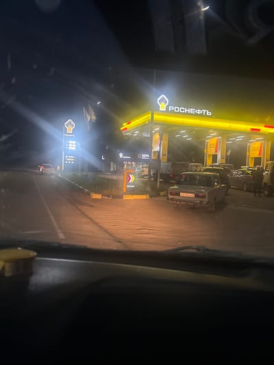 The image depicts a nighttime scene at a Rosneft gas station in the city of Petropavlovka, Russia. The gas station is brightly lit by yellow and white neon lights, with the Rosneft logo prominently displayed. Several cars are present, including a light-colored sedan at the entrance and multiple vehicles queued at the pumps. A truck is also visible. The ground appears to be asphalt, with some grass and a curb visible along the roadside. The overall atmosphere is functional and commercial, characteristic of a roadside service area at night.
