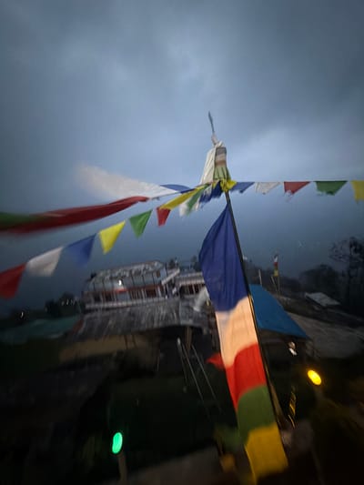 The image depicts a low-light scene in Ghandruk, Nepal, under an overcast, gray-blue sky. The foreground and midground are dominated by numerous Buddhist prayer flags, exhibiting motion blur consistent with wind or camera shake. A prominent vertical pole, positioned slightly right of center, displays a large, vertically striped flag in blue, white, red, green, and yellow. Above this pole, a horizontal string of smaller, multicolored prayer flags (red, yellow, green, white, blue) extends across the upper midground. The top of the central pole features a fabric-wrapped ornament, possibly culminating in a blade-like metallic object.
In the midground, behind the primary flags, a multi-story building with white walls and dark roofs is visible, displaying windows and balconies, with one interior light source illuminated. A substantial stone retaining wall is situated below the building. To the right of the central flag pole, a blue tarp or tent-like structure is present. The background shows indistinct, hazy hills or mountains obscured by mist or low clouds. Artificial light sources include a glowing green oval in the lower-left foreground and a circular yellow light in the lower-right. No individuals are discernible within the frame. The overall scene is characterized by dim illumination and motion blur across moving elements.