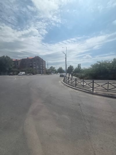 The image shows a street scene in the city of Border Gyumri-Akhuryan, Armenia. A multi-story building with a red roof is visible on the left side of the road. Several vehicles, including a truck, a white van, and a car, are parked near the building. A black metal fence runs along the right side of the road, separating it from a grassy area with trees. The sky is partly cloudy with patches of blue. The road appears to be asphalt with some visible cracks. Two people are walking on the right side of the road near the fence. There are also streetlights visible. The scene appears to be a typical urban environment.