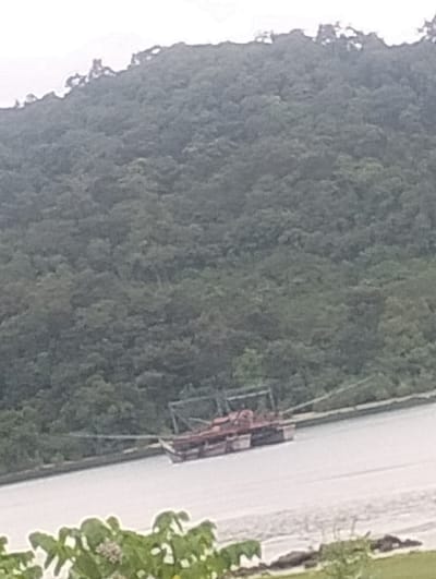 A wide shot shows a partially obscured view of a body of water with a boat on it. The boat appears to be a barge or a similar cargo vessel, with a red and white superstructure. It is connected to something off-frame by a thin line, possibly a tow rope. The water is a light, murky color, with some ripples on the surface. In the background, a densely forested hillside rises steeply, with the trees obscuring most of the terrain. The sky above is overcast and pale. In the foreground, the edge of a body of water is visible, with some green foliage and what appears to be rocks or debris. The location is specified as Layeun, Indonesia, though no specific landmarks or details confirm this in the image itself.