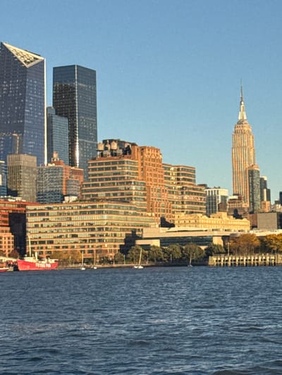 The image displays a daytime view of the Manhattan skyline from Weehawken, United States, looking east across the Hudson River under a clear blue sky. The foreground is occupied by the dark blue, rippled surface of the Hudson River. Along the Manhattan shoreline, a red-hulled lightship, labeled "FRYING PAN" in white, is prominently docked on the left. Several white-hulled sailboats with masts are moored in the river near the shore. A tree-lined promenade with some autumnal foliage is visible along the waterfront, alongside a wooden pier structure extending from the right.
The background is dominated by numerous high-rise buildings of the Manhattan skyline. On the left, a modern skyscraper with a distinctive angled, pointed top is visible, adjacent to other tall, dark glass-clad towers. Further right, the iconic Empire State Building, with its characteristic spire, stands out, illuminated in golden sunlight. Various other commercial and residential structures, ranging from multi-story buildings with stepped designs to conventional high-rises with glass, brick, and stone facades, form the urban density. Several rooftops feature cylindrical water towers. No individuals are discernible within the scene.