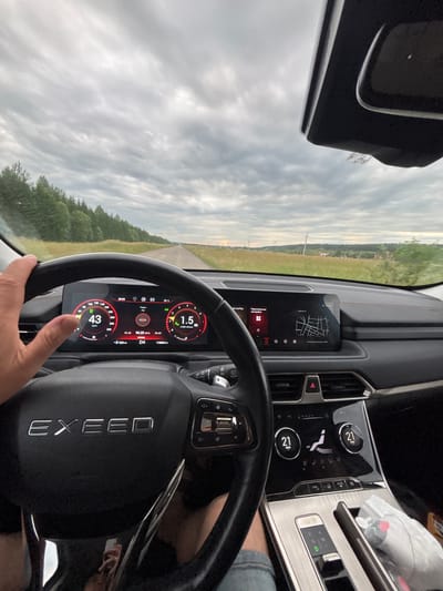 This is a point-of-view shot from inside a car, looking forward. The driver's left hand is visible on the steering wheel, which has the "EXCEED" logo. The car's dashboard displays various information, including speed (43 km/h), fuel consumption (1.5 l/100km), and temperature (19°C). The navigation screen is active, showing a map and the date 2020-06-19. The car is driving on a paved road through a rural landscape with trees and fields under a cloudy sky. The location is Kuregovo, Russia.