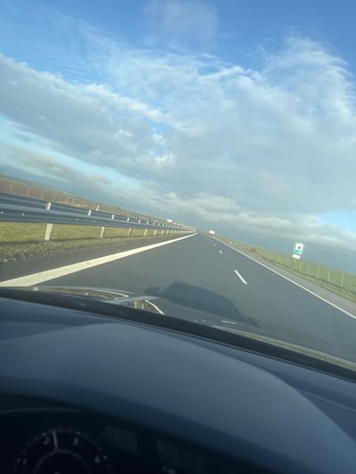 The image depicts a multi-lane asphalt highway, viewed from the interior of a vehicle, located in the vicinity of Vodenichane, Bulgaria. The sky is partially cloudy, featuring large white and grey cumulus clouds interspersed with patches of clear blue sky. The highway extends into the distance, marked with a solid white line on the left shoulder and a dashed white line indicating the right lane boundary.
To the left of the road, a metal guardrail with concrete posts separates the asphalt from a narrow grassy verge, beyond which lie brown and green agricultural fields extending to the horizon. To the right of the road, a similar grassy verge borders a wire fence. Past the fence, additional brown and green agricultural fields are visible. A rectangular road sign with a green border and a white center, containing indistinct green or black markings, is positioned on the right side of the road beyond the fence.
In the immediate foreground, the dark-colored dashboard of the vehicle is visible, with a portion of the steering wheel visible in the lower-left corner. A shadow from the vehicle is cast on the road surface. No individuals or other vehicles are distinctly visible within the frame. The scene is illuminated by natural daylight.
