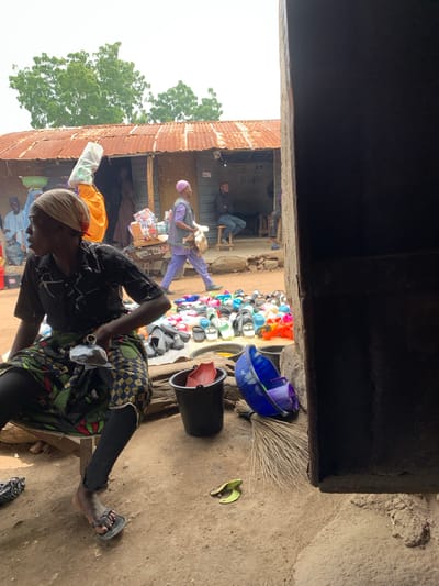 In the city of Dull A, Nigeria, a scene unfolds at an outdoor market stall. A woman, partially visible from the waist down, sits on a wooden bench wearing a patterned skirt and dark leggings, her right foot shod in a dark sandal. In front of her, a variety of colorful footwear is displayed on a white tarp. To her right, a black bucket contains a red fan, and a blue bucket and purple container are nearby, alongside a broom. The ground is sandy and uneven. In the background, a building with a corrugated metal roof stands, with people visible in and around it, some carrying goods. The overall impression is one of a bustling market environment.