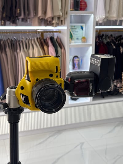 A yellow camera with a black lens and a black flash attachment is mounted on a tripod, with a blurred background of clothing racks and shelves. The camera and flash are the primary subjects, positioned in the foreground. The background suggests an indoor retail or studio environment, possibly a boutique or a photography studio. The location in Tanjungmeru, Indonesia is not discernable from the image.