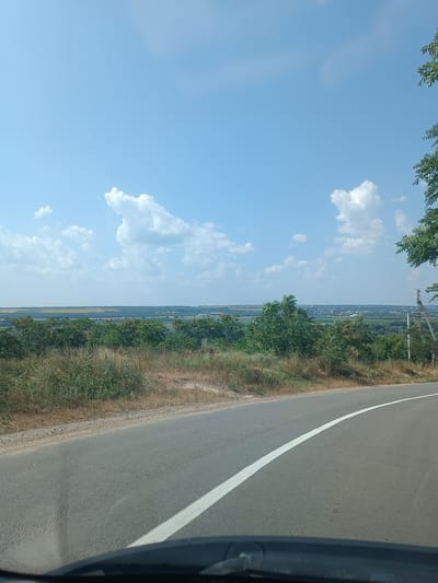 A wide-angle, eye-level, outdoor, daytime, full shot captures a scenic view of a rural landscape from the perspective of a vehicle, likely on a road. The foreground is dominated by a curved asphalt road with a white line marking its edge, disappearing into the distance. The road surface shows some cracks and wear.

The middle ground is characterized by a hillside covered with dry grass, dense green bushes, and scattered trees with reddish berries. A power pole with wires is visible on the right side of the image, slightly behind the vegetation.

Beyond the hillside, a vast panorama unfolds. A broad, flat plain stretches out, featuring agricultural fields of varying shades of green and yellow, interspersed with patches of trees and vegetation. In the far distance, a town or city is visible, with buildings and structures forming a faint outline against the horizon.

The sky is a clear, bright blue, with a few scattered white, puffy clouds. The overall impression is one of a sunny day in a tranquil, rural setting, potentially near Pohrebea, Moldova, given the landscape features. No people or animals are present in the frame.