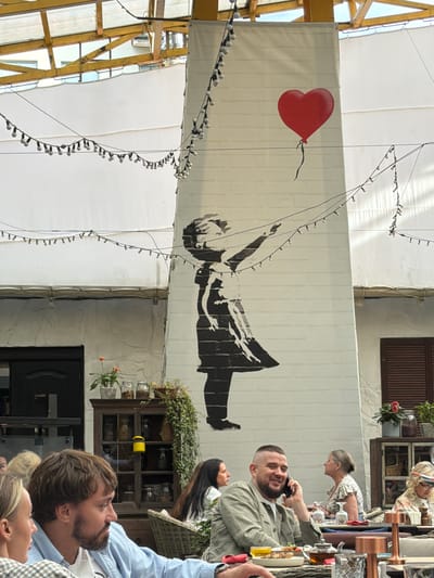 The image shows an outdoor seating area of a restaurant or cafe, likely located in Zhukovka, Russia. A large mural of a girl releasing a red heart-shaped balloon is painted on a white, pillar-like structure in the background. String lights with white bulbs are draped across the ceiling structure and around the mural. Several people are seated at tables, engaged in dining and conversation. In the foreground, a man with a beard and a light blue shirt is looking to his left, and a woman with blonde hair and a checkered shirt is partially visible on the far left. In the middle ground, a man in a green jacket is speaking on a cell phone and smiling. Other patrons are seated further back at their tables. The environment appears to be a covered outdoor space, possibly with a retractable roof or awning, suggested by the visible yellow-tinted ceiling structure. Decorative elements like plants in pots and antique-looking display cabinets are also present.