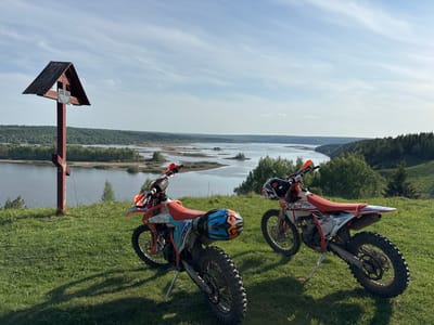 The image depicts a scenic overlook in Serga, Russia, featuring a wooden Orthodox cross structure on the left and two orange and white dirt bikes parked on a grassy hill. The bikes are facing the viewpoint overlooking a wide river interspersed with land formations and bordered by forested areas. The sky is bright with scattered clouds. No people are visible in the image.