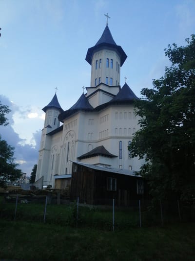 The photograph captures a white church with dark blue tiled roofs, featuring multiple spires topped with crosses. The church is illuminated by soft, natural light against a twilight sky with scattered clouds. In the foreground, a wooden structure with a tin roof is partially visible, and a metal fence encloses a grassy area. Dense green trees partially obscure the right side of the church. The scene is static and depicts an architectural structure, with no visible people or animals. The location is identified as the Fălticeni Monastery in Fălticeni, Romania.