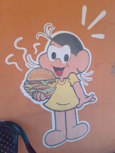 A cartoon illustration of a character resembling Monica from the Brazilian comic strip Monica's Gang is painted on an orange wall. The character is holding a large hamburger in its left hand and appears to be smiling. The illustration is outlined in white and black. White wavy lines emanate from the character's head, suggesting steam or excitement. To the right of the illustration, there is some faint graffiti or markings on the wall. In the bottom left corner of the image, a portion of a dark plastic chair is visible. The scene appears to be outdoors, likely on a building's exterior wall, in the city of São Gabriel Do Oeste, Brazil.