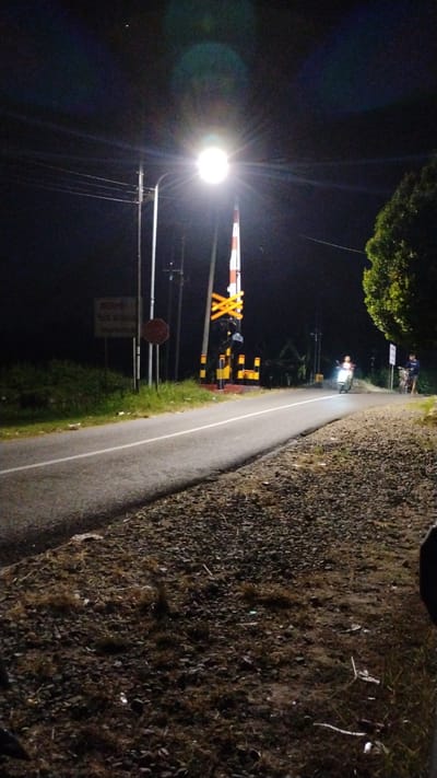 The image depicts a nighttime scene on a road near a railway crossing in Tulungrejo, Indonesia. A bright streetlamp illuminates the area, casting long shadows. A railway crossing signal, with alternating red and white bars and yellow crossbars, is prominent. Traffic lights are visible below the crossing signal. A red octagonal stop sign is mounted on a pole to the left of the road. A sign with text is also visible on the left side. The road has a white line marking its center. The shoulder of the road is covered in gravel and sparse vegetation. In the distance, a motorcycle with a rider is approaching the crossing. To the right, a person stands next to a bicycle. Lush green trees are visible on the right side of the image, and a dark, uncultivated area with grass and bushes lines the left side of the road. The sky is dark, indicative of nighttime, with some light glare from the streetlamp.