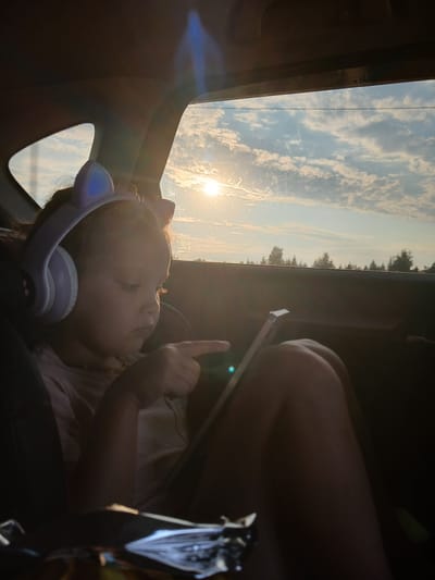 A young girl with cat-ear headphones is sitting in a car, looking at a tablet. The sun is setting outside the car window, casting a warm glow on the scene. The girl is wearing a pink shirt and a necklace. The car is likely in a rural area, as there are trees visible outside the window. The image appears to have been taken during a road trip. The location in Bol'shebadrakovo, Russia is not evident from the image.
