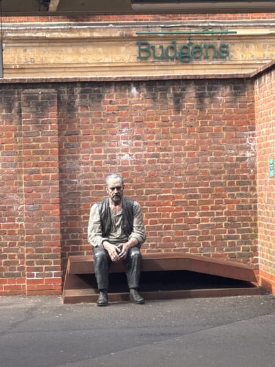 A weathered, life-sized statue of an elderly, bearded man is seated on a dark, angular metal bench in front of a red-brown brick wall. This scene is situated at Station Approach, Woking, United Kingdom, beneath a light-colored stone fascia displaying the green 'Budgens' retail sign. The statue, depicting a man in a shirt, waistcoat, and trousers with clasped hands, gazes forward with a somber expression. The brickwork shows areas of dark staining and light efflorescence. A small vertical blue-green sign with white numbering '3 11' is visible on the adjacent right wall section. The foreground consists of dark asphalt pavement.