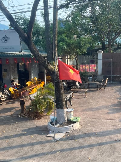 A street scene in M'Drắk, Đắk Lắk, Vietnam. A Vietnamese flag is displayed on a tree. Yellow chairs are stacked outside a building adorned with more Vietnamese flags. A child sits among the chairs. A parked motorbike is partially visible. A sign for "Trung Nguyên Legend" is also visible. The ground is paved with patterned tiles.