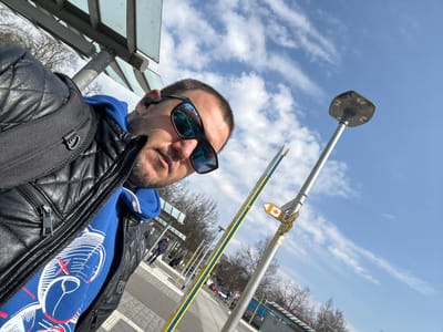 The image, captured in Überlingen, Germany, shows a male subject taking a selfie outdoors on a bright, partially cloudy day. The subject, in the foreground and tilted right, has short dark hair and a beard, wearing reflective blue sunglasses, a black quilted leather jacket, and a blue hooded sweatshirt with a white graphic design featuring red accents and X-shaped eyes. A black backpack is visible on his left shoulder. The background reveals an urban environment, likely a public transit stop. To the subject's right, a silver lamppost with a flat, square top stands beside a thinner silver pole and a yellow and blue striped pole, the latter featuring a small yellow and white directional sign. Above and behind the subject, the structural elements of a shelter, including metal framing and translucent panels, are visible. The ground is paved with light-colored material. Leafless deciduous trees and indistinct figures are present in the distant background.