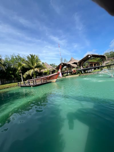 This media file depicts a tranquil, tropical waterside scene in Tambon Pa Kae Bo Hin, Thailand. The primary subjects include a wooden boat moored to a dock, several thatched-roof structures, lush greenery with palm trees, and a bright blue sky with scattered clouds. The water is a clear, vibrant turquoise, reflecting the surrounding environment.
The boat appears to be a traditional longtail boat, with its distinctive wooden hull and decorative elements. It is positioned alongside a rustic wooden pier that extends into the water. Behind the pier, the thatched-roof buildings suggest a resort or restaurant, with open-air designs characteristic of tropical architecture. The presence of a life preserver ring attached to a railing on one of the structures implies a focus on safety and water activities.
The scene is bathed in bright sunlight, indicating it is daytime, likely mid-morning or afternoon, given the clear sky and the quality of light. The weather appears to be warm and pleasant, typical of a tropical climate.
Notable details include the reflection of a person in the water, possibly the photographer, which adds a personal element to the otherwise scenic view. The overall emotion conveyed is one of peace and relaxation, inviting viewers to imagine themselves in this idyllic setting. The location cues, such as the specific type of vegetation and architecture, strongly suggest a Southeast Asian tropical environment, specifically Thailand as indicated by the provided location context. No visible text is present in the image.