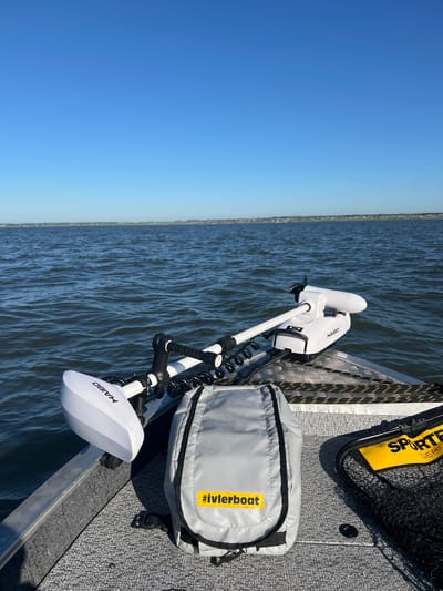 A perspective shot from the front of a boat on a body of water under a clear blue sky. The boat's bow, made of gray textured material, is visible in the foreground, with a metal railing along the edge. Mounted on the boat is a white electric trolling motor with the brand name "HAIBO" visible on its housing. Next to the motor is a gray bag with a yellow tag that reads "#ivlerboat". To the right of the bag is a black fishing net with a yellow handle labeled "SPORTEX GERMANY". In the background, a calm body of water stretches to the horizon, where a distant shoreline with trees and houses can be seen. The location is the city of Novopichugovo, Russia.