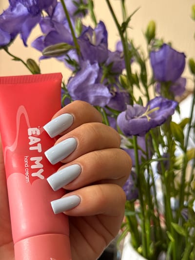 A close-up shot shows a person's hand with light blue-painted nails holding a pink tube of "EAT MY hand cream". The hand is positioned in front of a bouquet of purple delphinium flowers, some of which are in bloom and others are buds. The flowers are a vibrant purple and green, with delicate petals and long stems. The background is softly blurred, suggesting a shallow depth of field. The overall lighting is natural and diffused, creating a soft glow on the hand and flowers. The image has a gentle and feminine feel, possibly evoking a sense of self-care or beauty. The specific location context of Verkhnii Lars, Russia, does not appear to be visually represented in the image itself, but can be assumed as the setting for this still life. The text on the cream tube, "EAT MY hand cream", is clearly legible.
