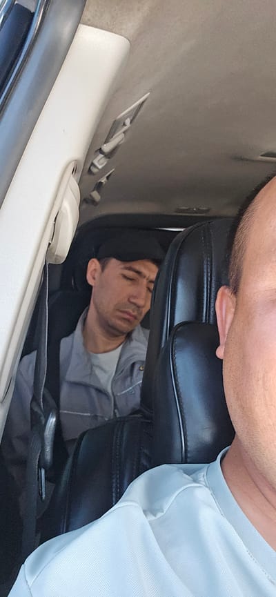 In the city of Guzar, Uzbekistan, a man wearing a grey work uniform and a black baseball cap is asleep in the back seat of a vehicle. The man is slumped against a dark-colored headrest, his eyes closed and his mouth slightly open. The interior of the car is visible, including a white door panel on the left and black leather seats. In the foreground, the shoulder and neck of another person, likely the photographer, are visible, wearing a light blue shirt. The scene appears to be a daytime interior shot.