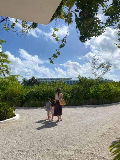 The image depicts an outdoor scene with a woman and two children walking along a gravel path in Marina Maroma, Mexico. Lush green vegetation lines the path, and a multi-story building is visible in the background amidst palm trees. The sky is blue with fluffy white clouds. The perspective is slightly angled upwards, capturing trees and foliage framing the sky.