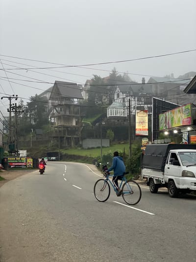 The image depicts a street scene in Seetha Eliya, Sri Lanka, characterized by a misty, overcast day. A cyclist wearing a blue jacket is riding a light blue bicycle on the right side of the road. A white truck is parked beside the cyclist, and a motorcycle with its taillight illuminated is positioned further down the road.
Along the left side of the road, there is a building under construction and various commercial advertisements, including signs for "Afra Grocery" and "KARUNANAYAKA TYRE HOUSE," as well as power lines running overhead. The environment is lush with greenery, indicating a mountainous region.