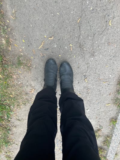 The image shows a person's legs and feet standing on a cracked concrete surface in Cherliany, Ukraine. The person is wearing black pants and black leather motorcycle boots. The concrete is light gray and has small cracks and patches of discoloration. There are scattered dead twigs and dry grass visible on the concrete and along the edges of the frame. A patch of green grass is visible along the top and right edges of the frame.
