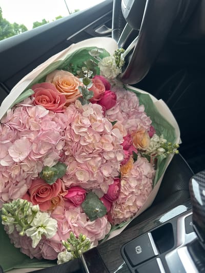 A bouquet of pink hydrangeas and roses is displayed in the passenger seat of a car. The bouquet is wrapped in green paper and white ribbon. The car's interior is visible, including the steering wheel and dashboard. The scene is likely in Hoffman Estates, United States of America, as indicated by the provided context.