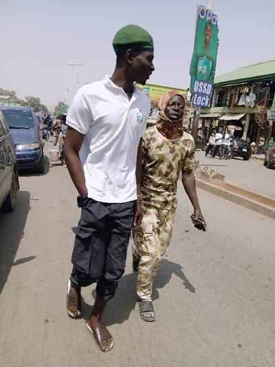 Two individuals walk on a paved street under clear, bright skies. On the left, a tall male wears a green beret, a white short-sleeved polo shirt with a small green and white emblem on the left chest, dark grey or black three-quarter length cargo pants, and brown open-toe sandals. His left hand is partially in his pocket, and he looks to his right. To his right, a shorter individual, presenting as an older adult, wears a camouflage pattern (tan and green) long-sleeved shirt and pants, a brown and tan patterned head covering that exposes the face, and dark grey open-toe sandals. This individual holds a small, light-colored object in their right hand and looks generally forward.
Behind the two main subjects, on the right side of the street, a tall vertical billboard stands, displaying the text "OPA" at the top, a smiling male face, and text including "#" and "USSD Lock". Further into the background, a multi-story building with visible windows and hanging items is present. A dark blue minivan or similar vehicle is partially visible on the far left, in the background. Other indistinct figures are scattered throughout the mid-ground and background, suggesting a public, populated urban area. Shadows of the main subjects are cast on the road, indicating strong sunlight.
