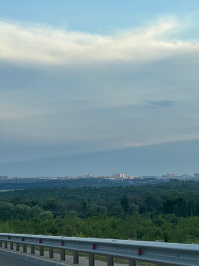 The image depicts a panoramic view of a city skyline in Novomar'evskaya, Russia, seen from a slightly elevated vantage point. In the foreground, a silver metal guardrail with red reflectors runs horizontally across the lower portion of the frame, suggesting the perspective is from a road or elevated pathway. Beyond the guardrail, a dense expanse of lush green trees covers the mid-ground, forming a natural buffer between the viewer and the urban landscape.
The city skyline, located in the distance, is characterized by a collection of multi-story buildings, including apartment blocks and other urban structures. The buildings are illuminated by soft, warm light, indicating either dawn or dusk. A tall, slender structure, possibly a radio tower or a communication mast, stands prominently amongst the cityscape.
The sky above is a mix of pale blue and diffused white clouds. A large, bright white cloud formation stretches across the upper portion of the image, with lighter wisps and streaks of white scattering across the pale blue expanse. A faint, horizontal band of shadow or darker sky is visible in the mid-sky, possibly cast by a cloud formation not entirely within the frame. The overall atmosphere of the image is serene and suggests a peaceful, early morning or late evening observation of the city. There are no people or visible animals in the frame.